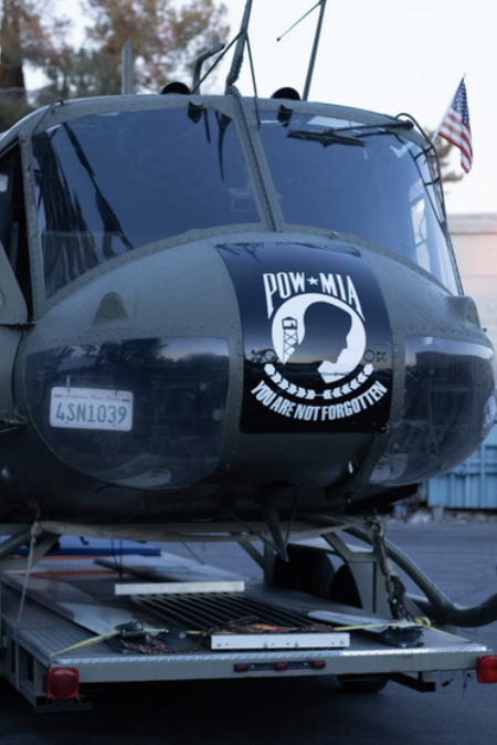 A military helicopter parked on a trailer, displaying a POW-MIA sticker with a silhouette of a soldier and the message "You Are Not Forgotten," and an American flag in the background.