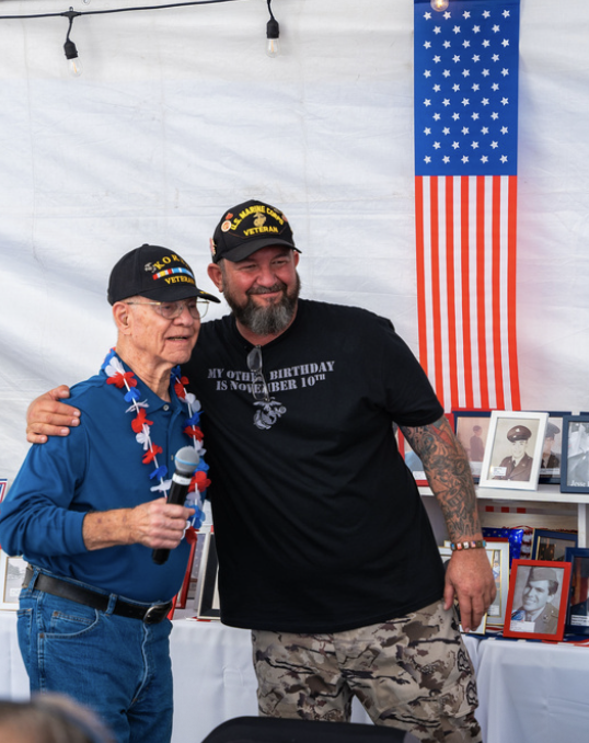 Two men, one elderly and one middle-aged, standing together at a patriotic celebration, with American flag-themed decorations and photos in the background.