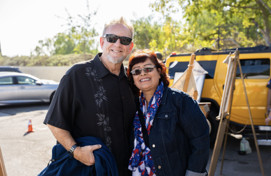A smiling man and woman standing outdoors with trees in the background, wearing sunglasses, and posing for the photo.