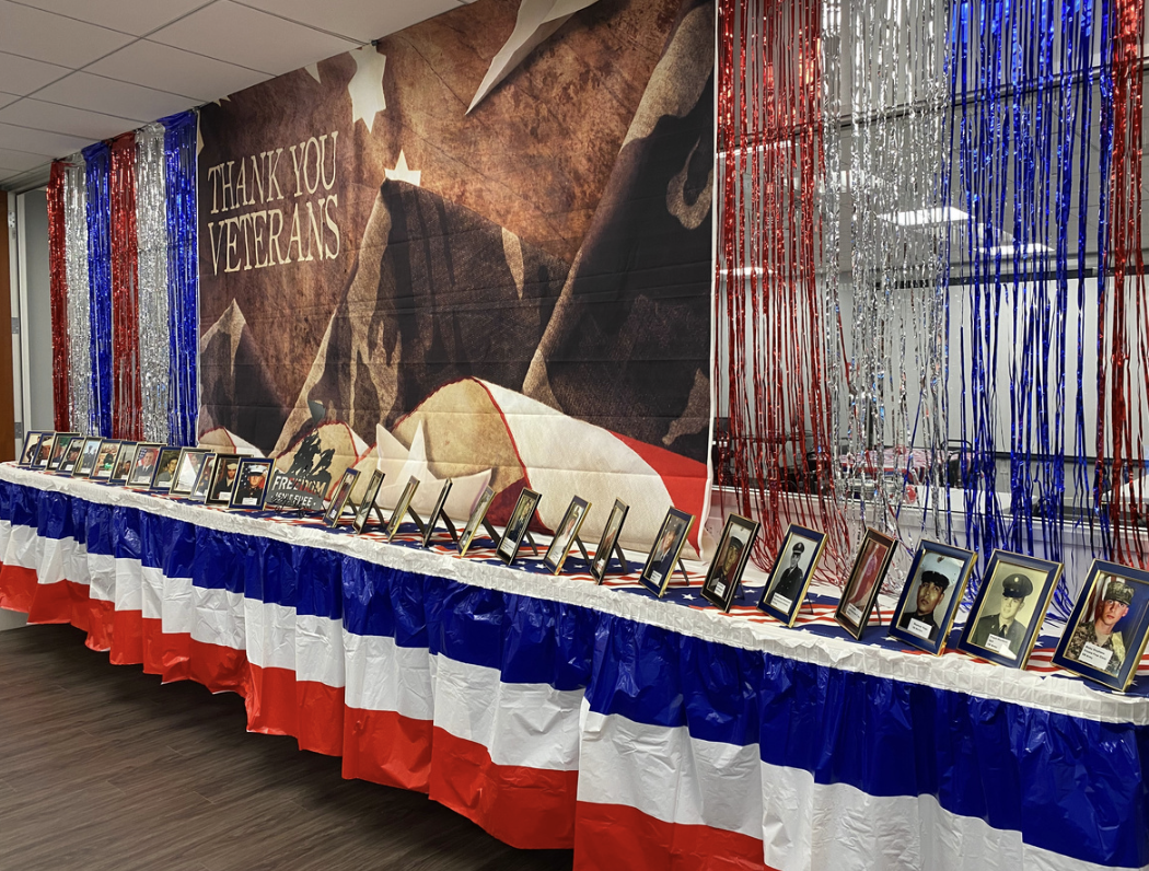 A display honoring veterans with framed photographs of military personnel on a table draped with a patriotic tablecloth. The background features a mural of an American flag and a message saying "Thank You Veterans." Red, white, and blue streamers and