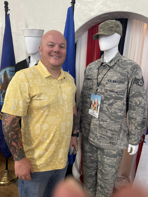 A man with tattoos wearing a yellow Hawaiian shirt standing next to a mannequin dressed in a U.S. Air Force camouflage uniform, with a cap and an ID badge, in front of a white wall and flags.