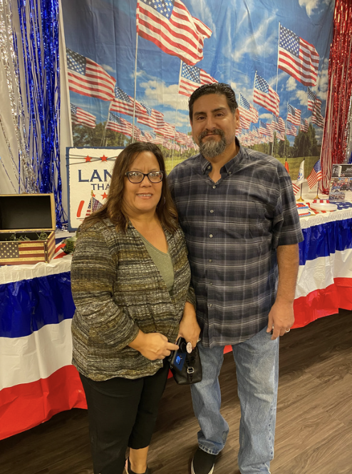 A woman and a man standing side by side in front of a patriotic display with American flags and red, white, and blue decorations. The woman is wearing glasses and a patterned blazer, while the man has a beard and is wearing a plaid shirt.