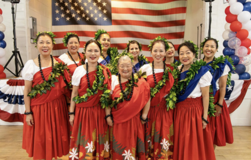 Group of women in traditional Hawaiian attire standing in front of American flag and patriotic decorations.