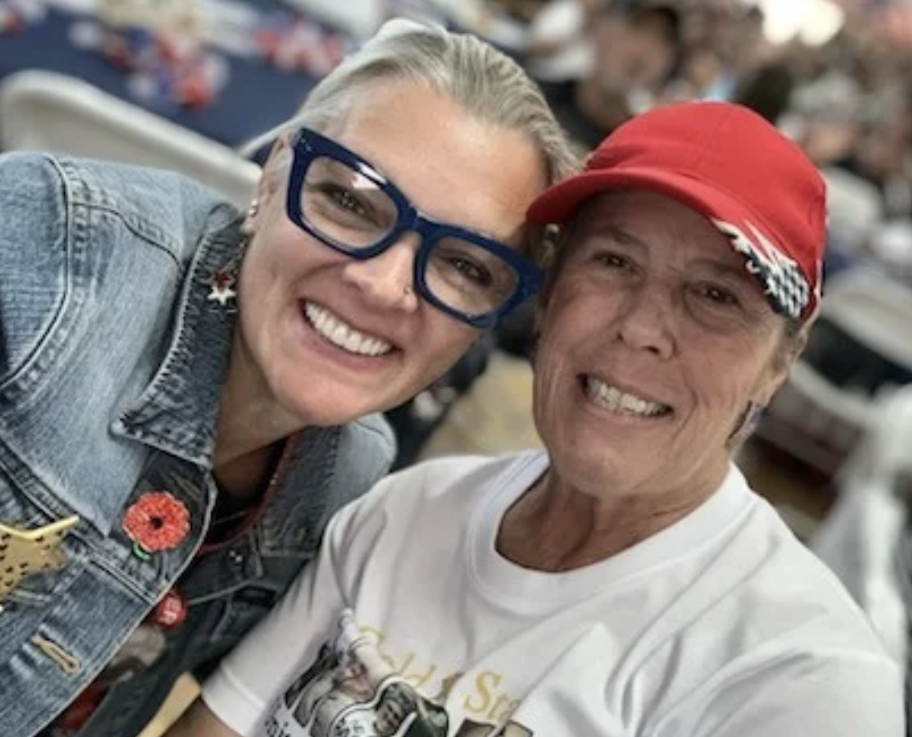 Two smiling women take a selfie together. One woman has blonde hair, wears glasses, a denim jacket with pins, and flower earrings. The other woman has short hair, wears a red cap, and a white T-shirt. They are smiling with a blurred background of a crowd and cars.