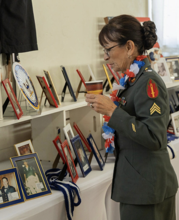 A woman in a military uniform with medals and an American flag-themed lei, looking at framed photographs and memorabilia on a display shelf.