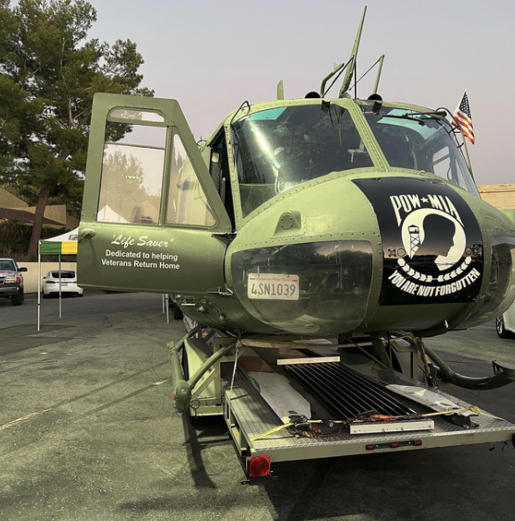 Military helicopter on a trailer in a parking lot, with a POW/MIA emblem and an American flag, dedicated to helping veterans return home.