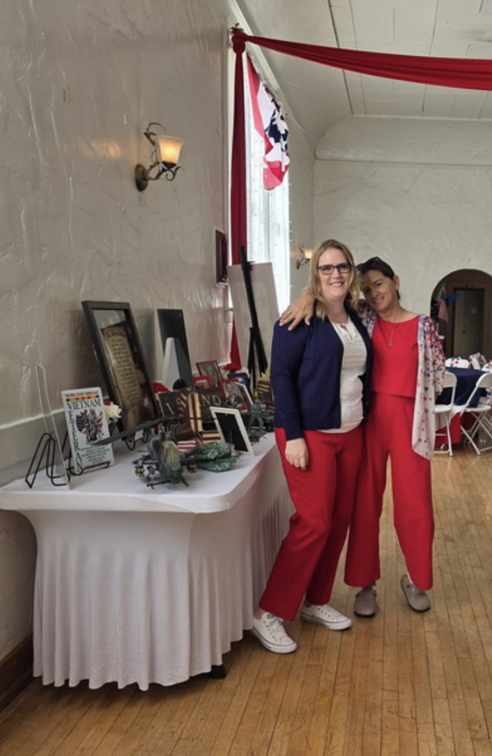 Two women standing together with one arm around each other, smiling. They are in a decorated event space with a table of framed photos and memorabilia nearby, and patriotic red, white, and blue decorations in the background.