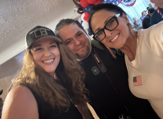 Three people smiling for a selfie at a celebration or event with American flag decorations and red, white, and blue balloons in the background.