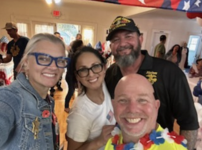 Four people smiling and posing for a photo at a celebration or gathering, with patriotic decorations in the background.