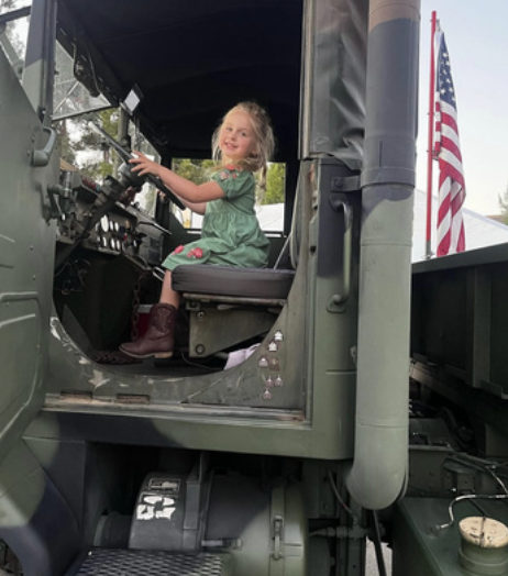 A young girl sitting in the driver's seat of a military vehicle with an American flag in the background.