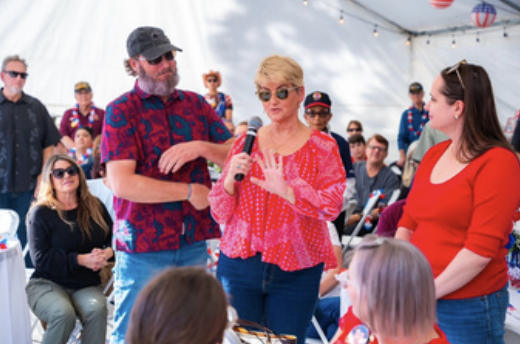 A woman speaking into a microphone at a gathering inside a tent, with people seated and standing around her.