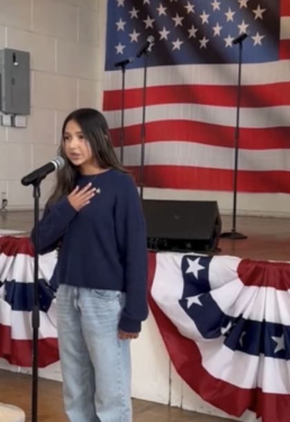 Young girl speaking into a microphone on a stage decorated with American Flags at a patriotic event.