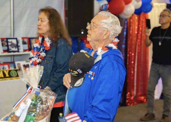 Two elderly veterans wearing patriotic leis and jackets, standing at an event decorated with red, white, and blue balloons and curtains, with one holding a cap with medals.
