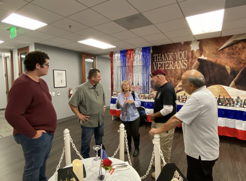 Group of people talking in a room decorated for Veterans Day, with a large 'Thank You Veterans' banner and photos on a table.