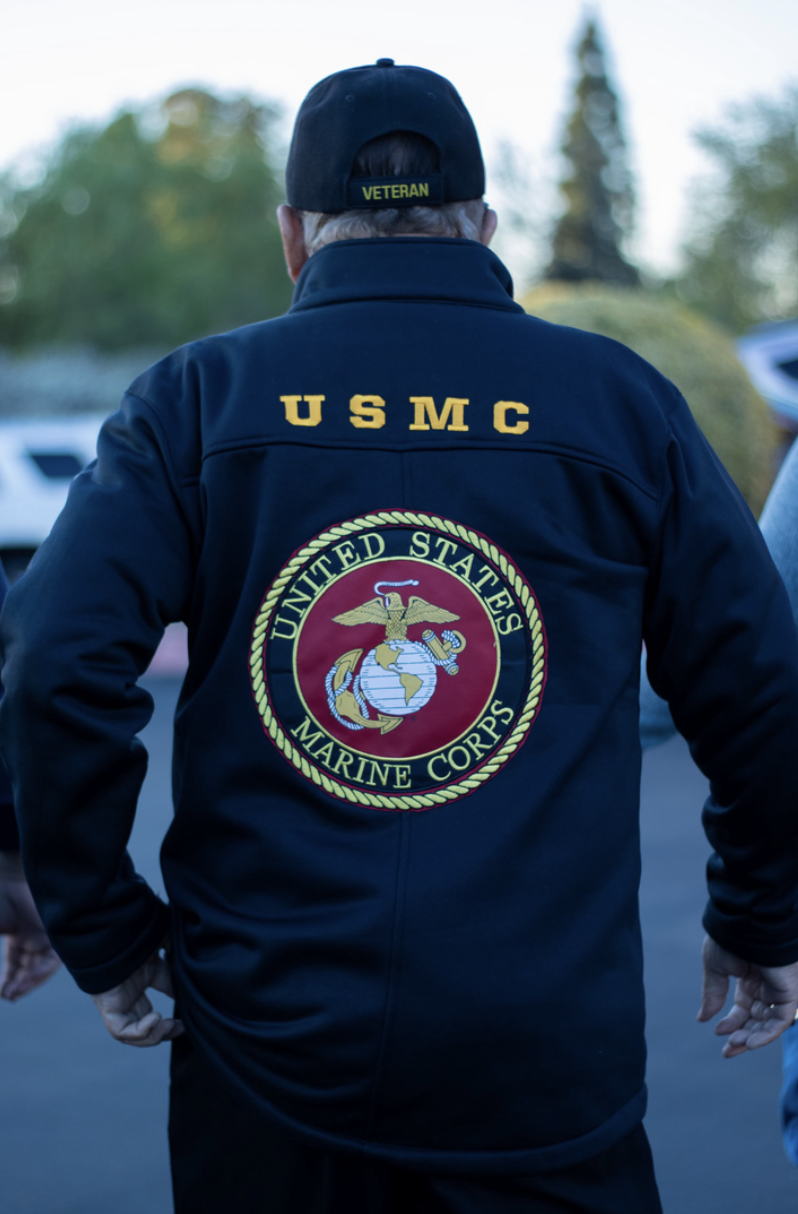 Back view of a U.S. Marine Corps veteran wearing a navy jacket with the Marine Corps emblem and the inscription "USMC" on the back, along with a black cap labeled "Veteran".