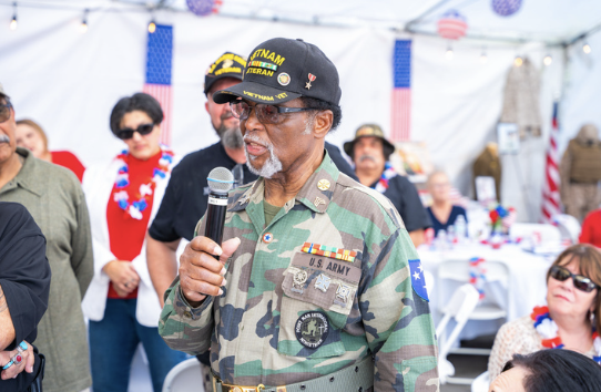 An elderly man in U.S. Army camouflage uniform, wearing a black cap, speaking into a microphone at a patriotic outdoor event, with people dressed in red, white, and blue in the background.