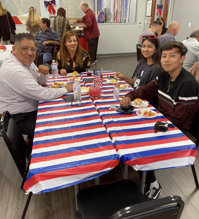 People sitting at a table with a red, white, and blue striped tablecloth, eating food and drinking water bottles, with holiday decorations in the background.