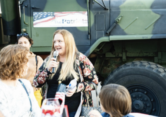 A woman with long blonde hair speaking into a microphone at a patriotic event, standing in front of a military vehicle with a flag-themed banner, surrounded by several people.