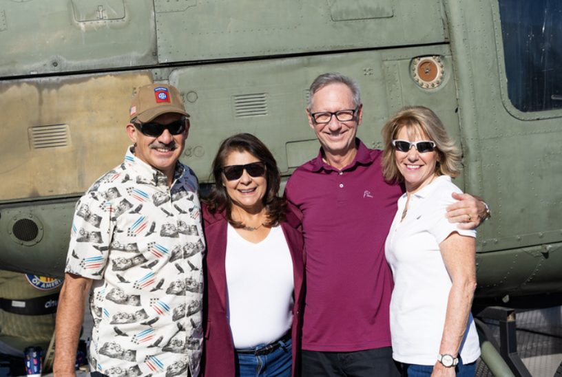Four smiling adults standing close together in front of a military helicopter, outdoors, wearing casual clothing and sunglasses.