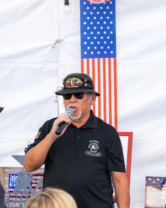 A man in a black US Army shirt and sunglasses speaking into a microphone in front of a patriotic American flag and a white tent.
