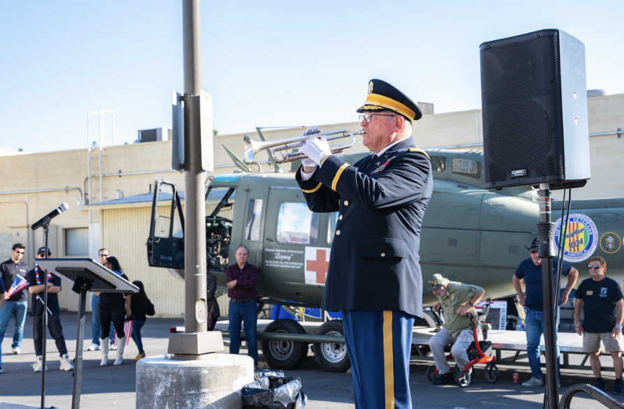 An elderly man in a military uniform with a cap playing a trumpet at an outdoor event, with a helicopter and several people in the background.
