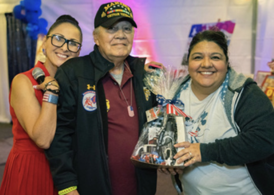 Three people smiling, with one holding a gift basket, in a decorated indoor setting