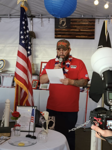 A man wearing a red shirt and a USA flag lei, reading from a paper at a patriotic event. He has a beard, glasses, tattoos, and a camo cap with red letters that says 'USMC'. An American flag stands next to him. The background features patriotic decora