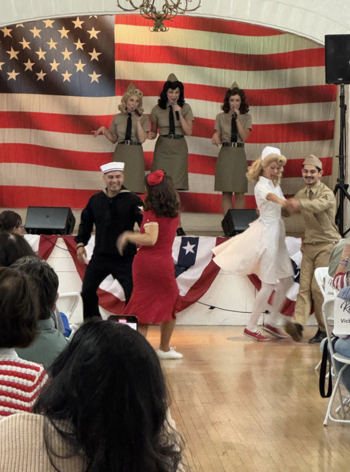 People performing a dance on stage, with three women in military-style uniforms singing in the background, during an event celebrating American patriotism with an American flag backdrop.