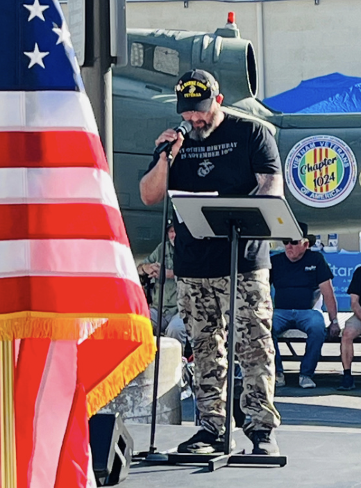 Man wearing a black hat and camouflage pants speaking into a microphone at an outdoor event, with an American flag in the foreground and a military helicopter in the background.