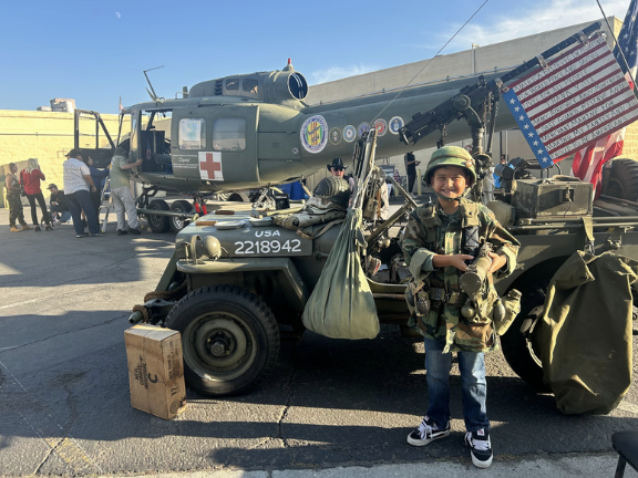 A young boy in military gear standing next to a military vehicle with an American flag and a helicopter in the background.
