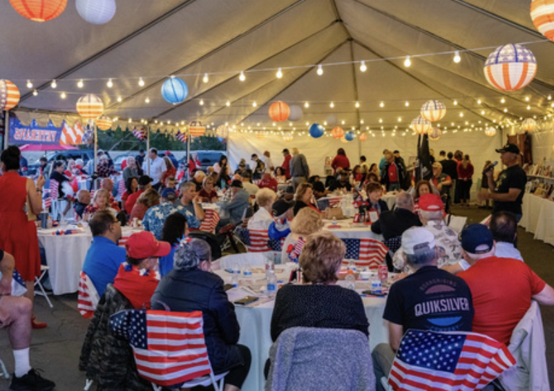 A decorated outdoor gathering with people under a tent, featuring American flags, red, white, and blue theme, hanging lanterns, and string lights.