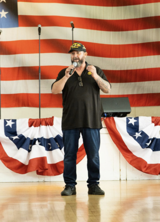 A man with a beard and tattoos, wearing a black shirt and cap, speaking into a microphone on a stage decorated with American flag-themed bunting and a large American flag backdrop.