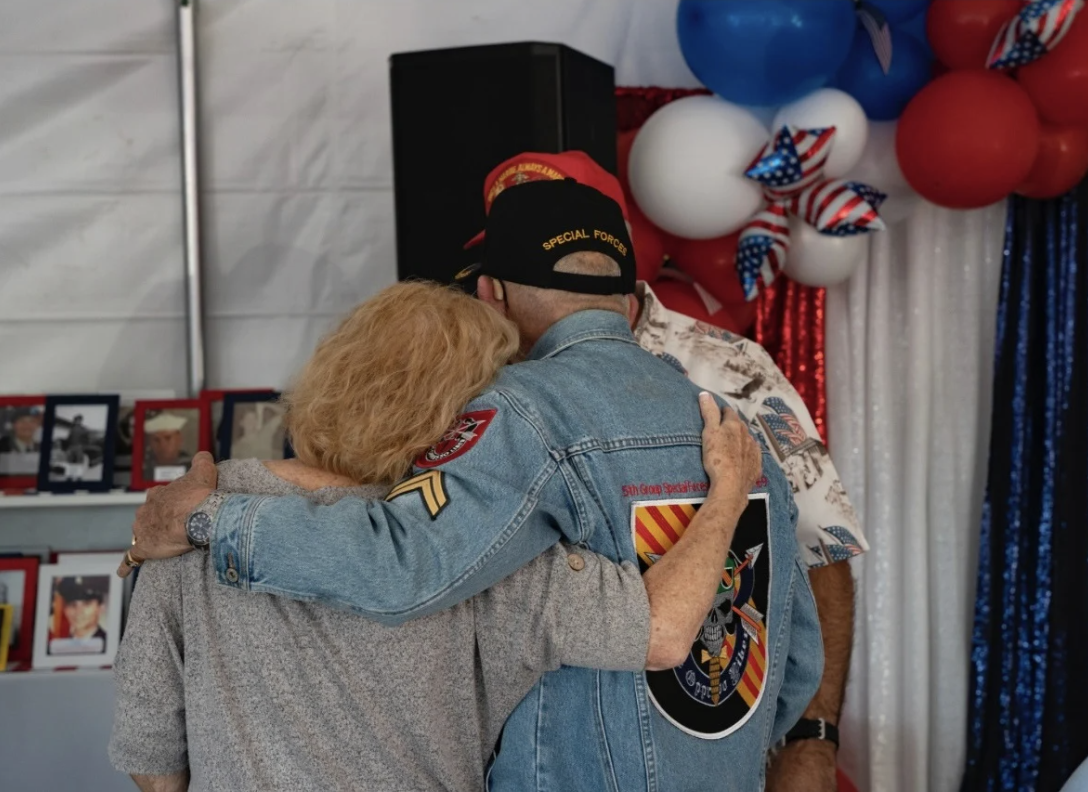Three people embracing at a patriotic event with balloons and photo display in the background, one man wearing a Special Forces cap and denim jacket.