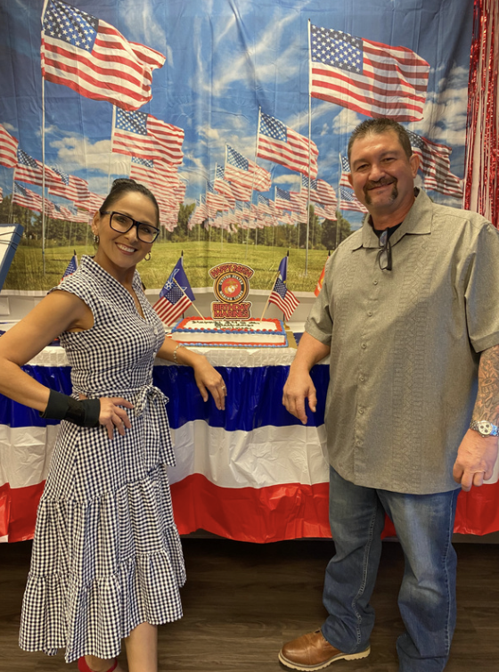 A woman and a man standing in front of a table with a patriotic American flag backdrop, a cake, and small American flags. The woman is wearing a checkered dress and glasses, and the man is wearing a gray shirt and jeans. The setting appears to be a c
