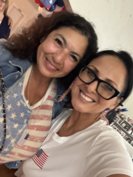 Two women smiling for a selfie, celebrating with patriotic clothing and accessories, in an indoor setting with patriotic decorations.
