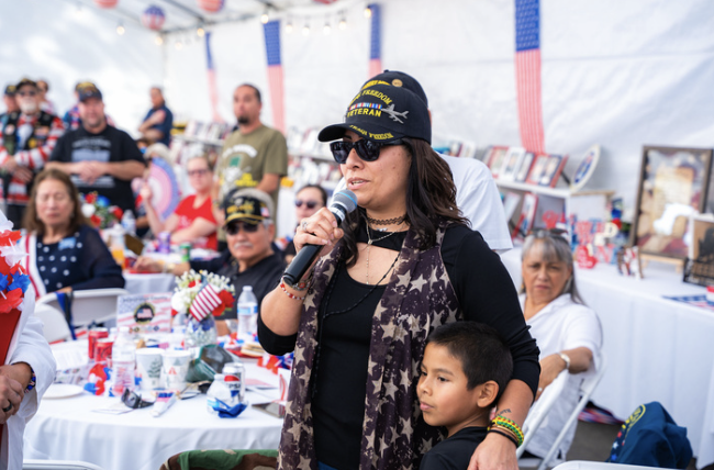 A woman wearing a black cap, sunglasses, and a camouflage scarf speaking into a microphone at a patriotic event with a group of people behind her inside a decorated tent.