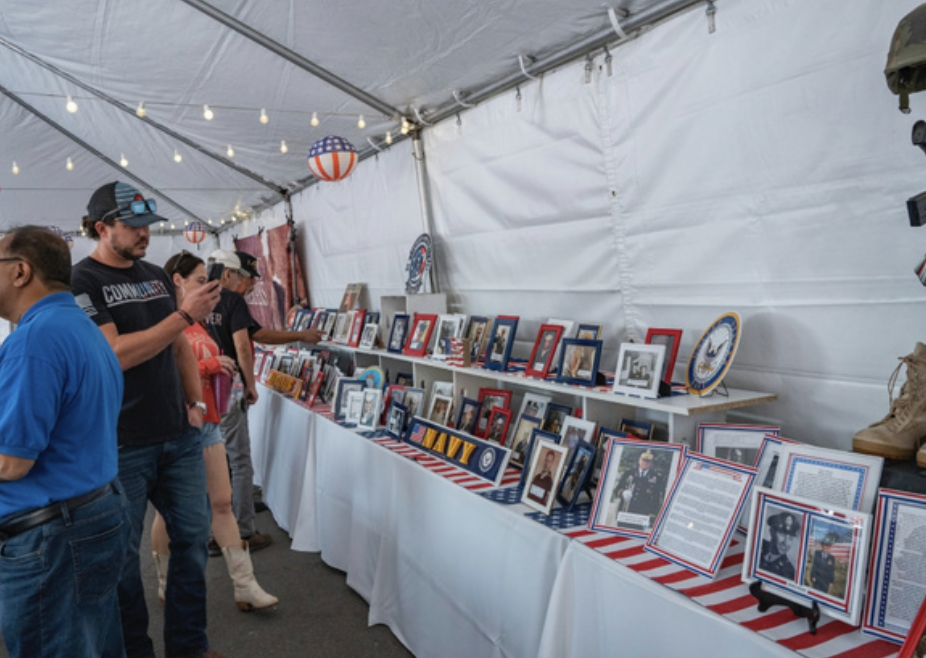 People browsing framed photographs and memorabilia at a display table inside a tent with patriotic decorations and military-themed items.