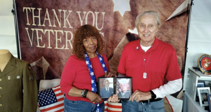 An elderly man and woman stand together holding photos, in front of a Veterans Day display that says "Thank You Veterans." The woman has curly hair and is wearing a red shirt with a star-spangled scarf, while the man has white hair, a red shirt, and 