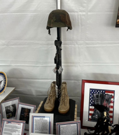 Military helmet placed on a rifle, which is mounted on a pair of tan combat boots, displayed on a black stand. Surrounding them are framed photographs and documents.