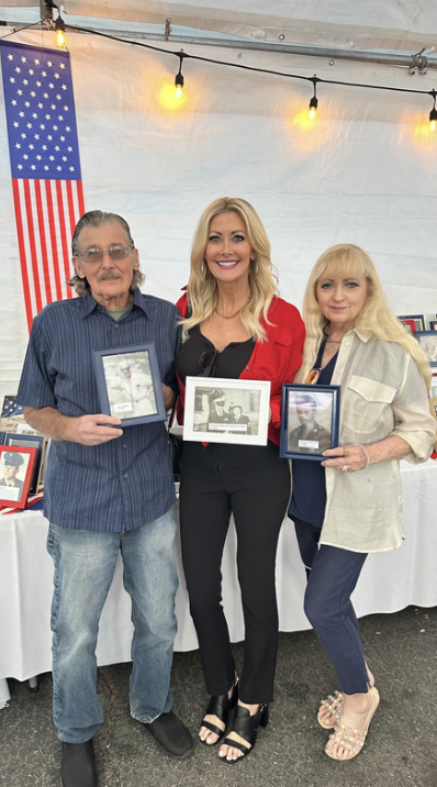 Three women standing together, holding framed photos, outdoors under string lights, with an American flag in the background.