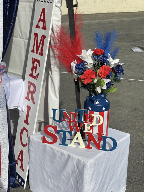 A table with patriotic decorations, including a tall patriotic-themed vase with red, white, and blue feathers and floral arrangement, a sign that says 'America', and colorful letters spelling 'United we stand' in red, white, and blue.
