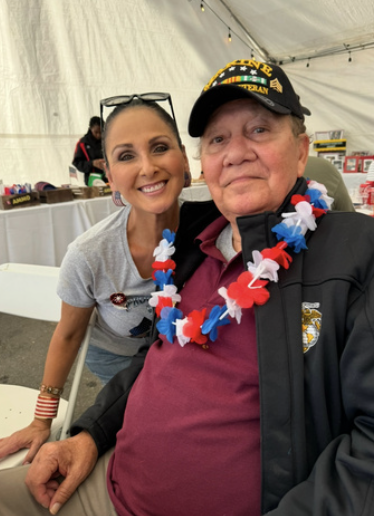 A woman and an older man taking a selfie at an event. The woman has dark hair, sunglasses on her head, and is smiling. The man is wearing a black cap with yellow text, a black jacket, and a red, white, and blue flower lei. They are inside a tent with