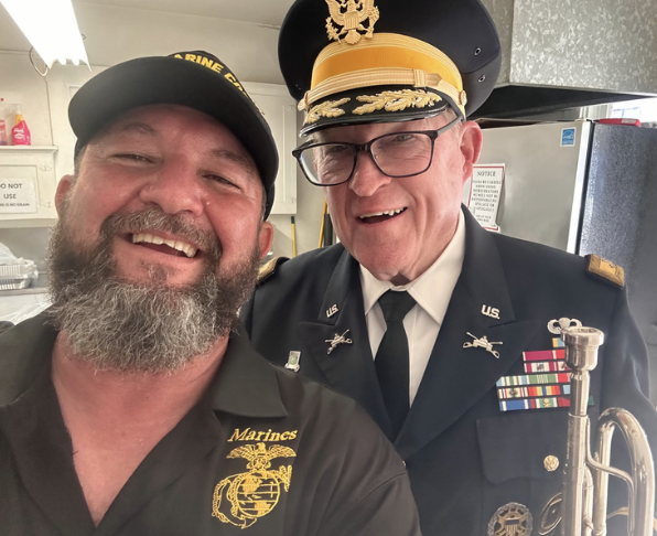 Two men smiling, one in a U.S. Marine Corps uniform and the other in a Marine veteran shirt, taking a selfie together. The man in uniform is wearing a peaked cap and glasses, and the other man is wearing a baseball cap. They are indoors, likely a kit