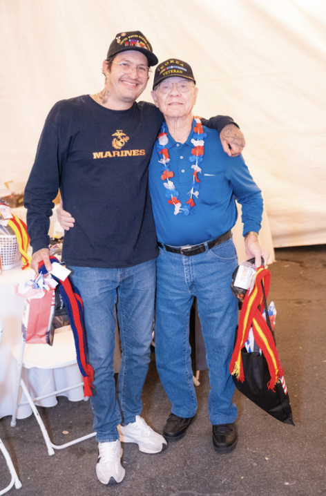 Two men standing together, smiling, with patriotic decorations. One is younger, wearing a Marine Corps shirt and a cap, the other elderly, wearing a blue shirt, a patriotic lei, and a veteran cap. They are holding bags with red, yellow, and black rib