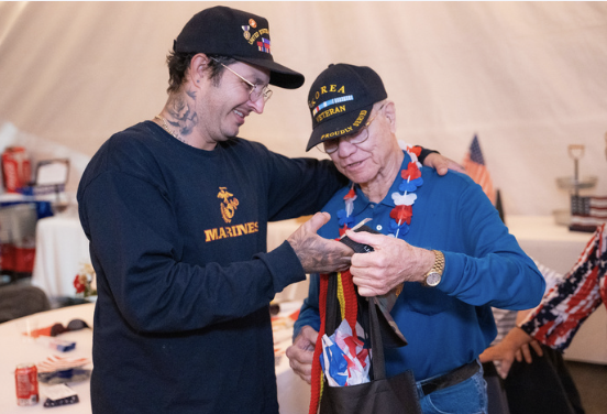 Two men, one in a Marines sweatshirt and the other in a blue shirt with patriotic accessories, look at a smartphone together inside a tent, exchanging smiles.