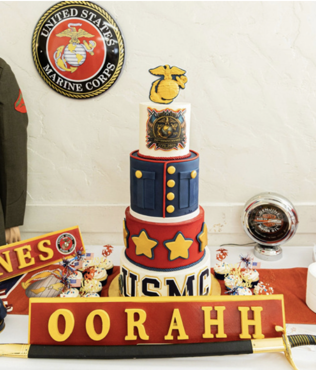 A four-tiered cake with a US Marine Corps theme. The top tier has an inflatable Marine emblem. The second tier looks like a Marine uniform with yellow buttons. The third tier is red with yellow stars. The bottom tier has the letters "USMC". The cake 