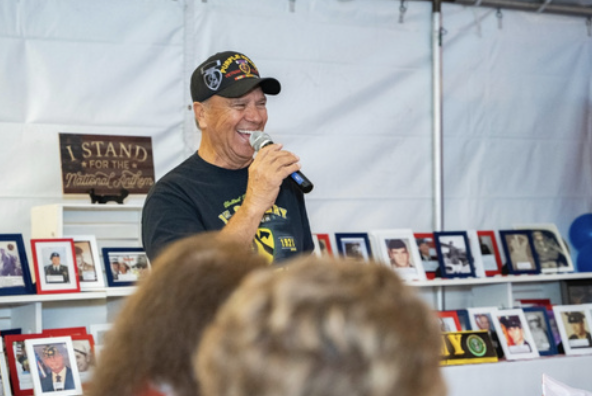 A man wearing a black cap and navy T-shirt speaking into a microphone at a gathering with photo displays behind him.