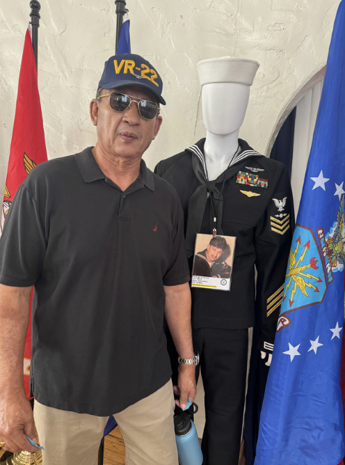 Man wearing sunglasses and a VR-22 cap standing next to a mannequin dressed in U.S. Navy uniform, with flags in the background.