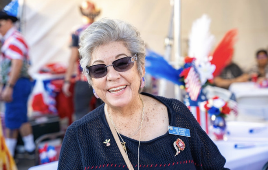 Smiling elderly woman with short gray hair wearing sunglasses and patriotic jewelry at a festive outdoor event decorated with red, white, and blue decorations.