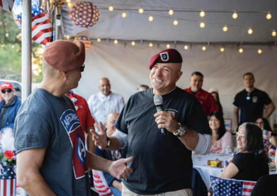 A man in a black T-shirt and red beret smiling and speaking into a microphone, interacting with another man in a blue T-shirt and glasses at a patriotic celebration party. People are seated at tables decorated with American flags, bunting, and string
